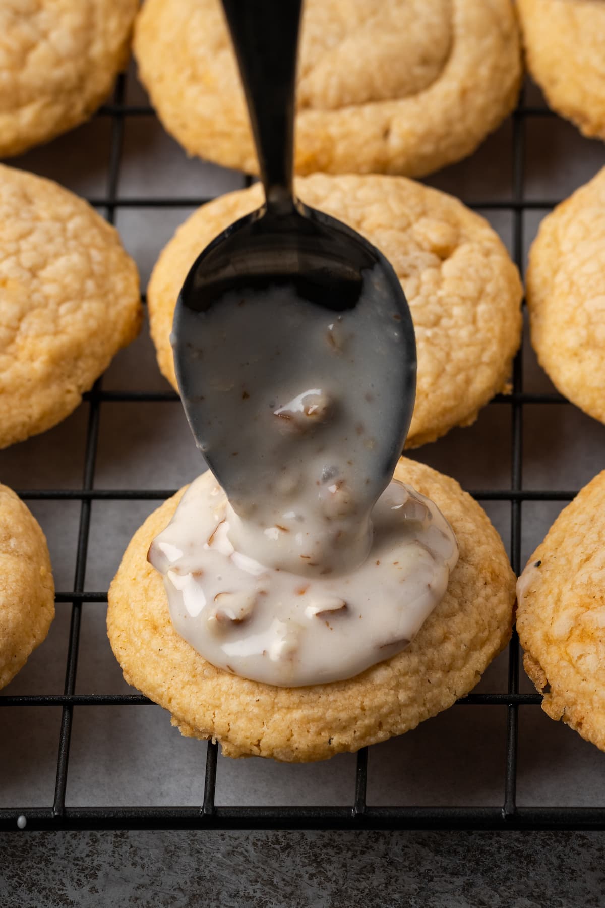 A spoon drops glaze on top of a butter pecan cookie on a wire rack.