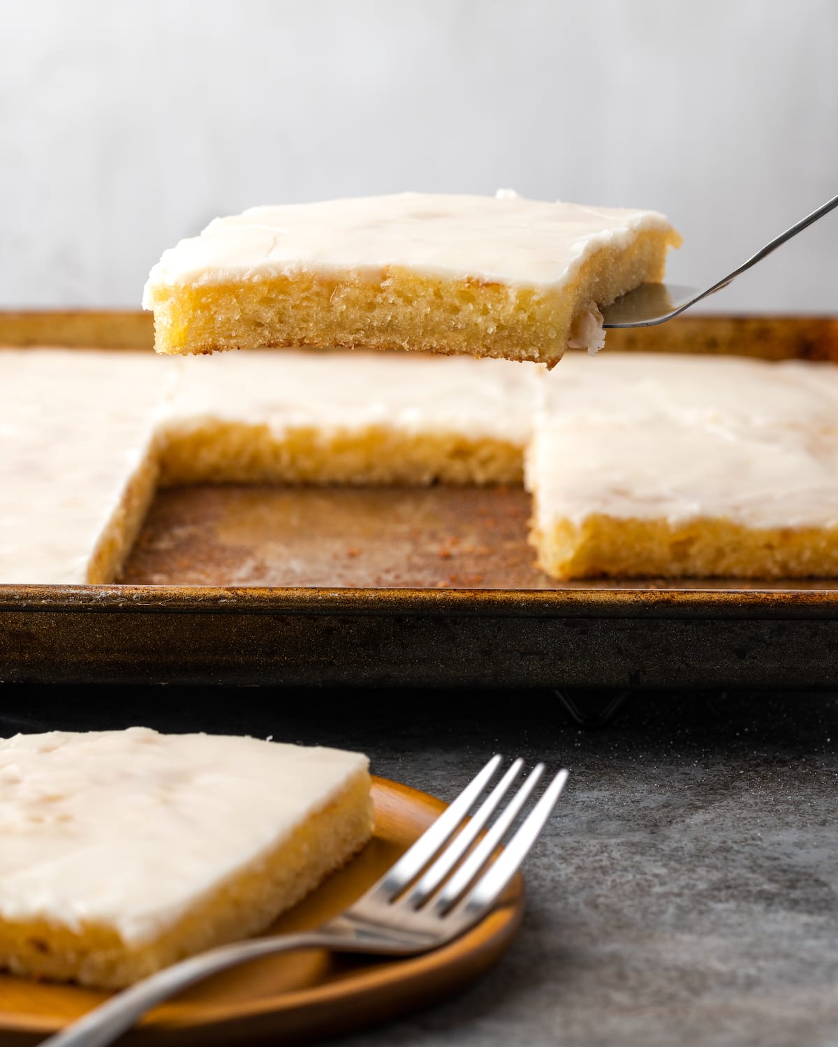 A slice of buttermilk sheet cake is lifted from a pan, next to a slice of cake on a plate with a fork.