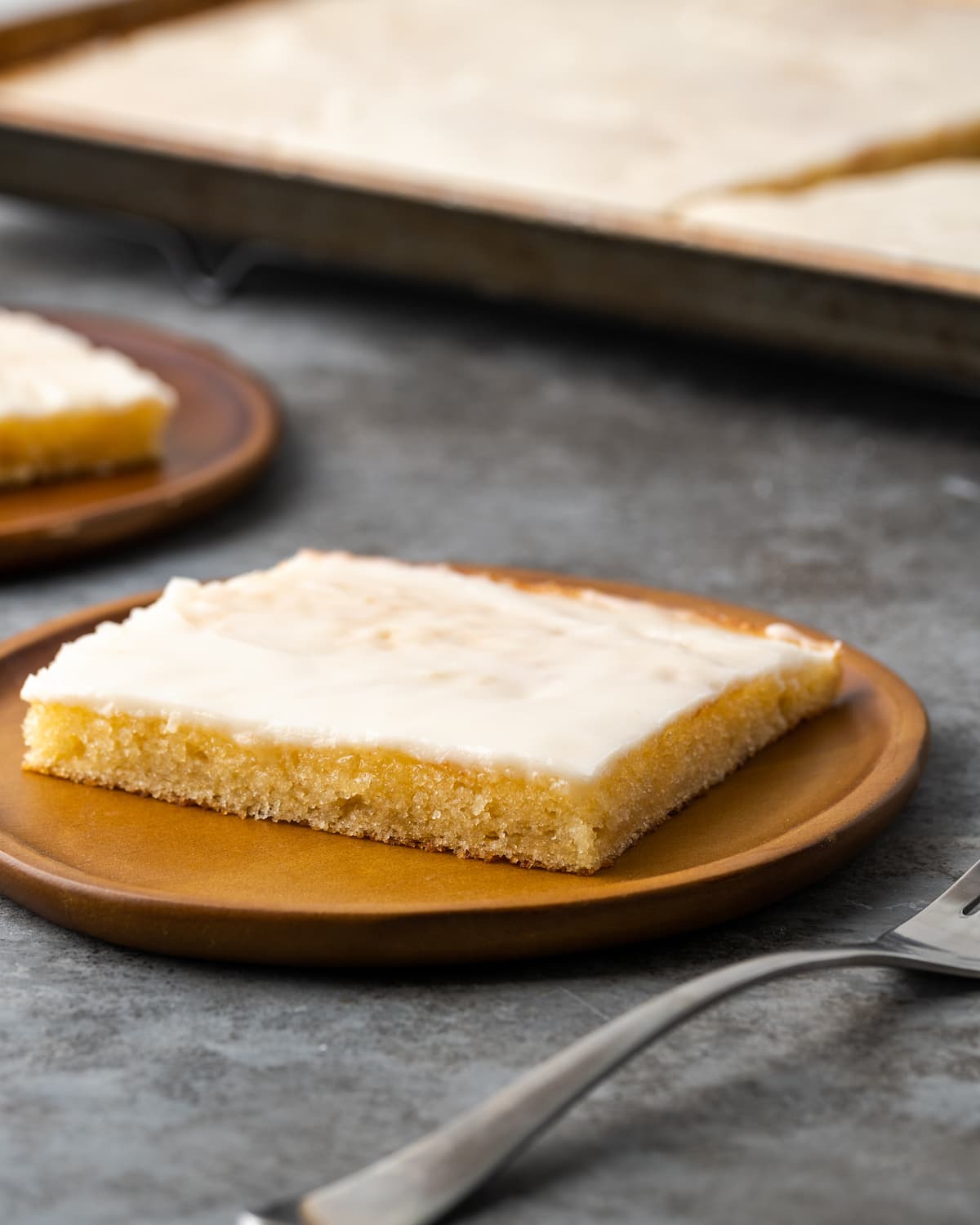 A slice of buttermilk sheet cake on a brown plate next to a fork.