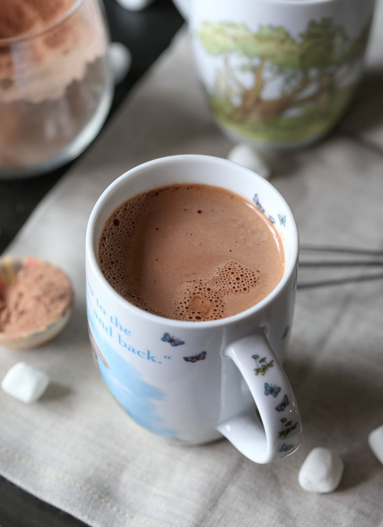 A mug of homemade hot chocolate next to scattered mini marshmallows and a jar of hot cocoa mix.