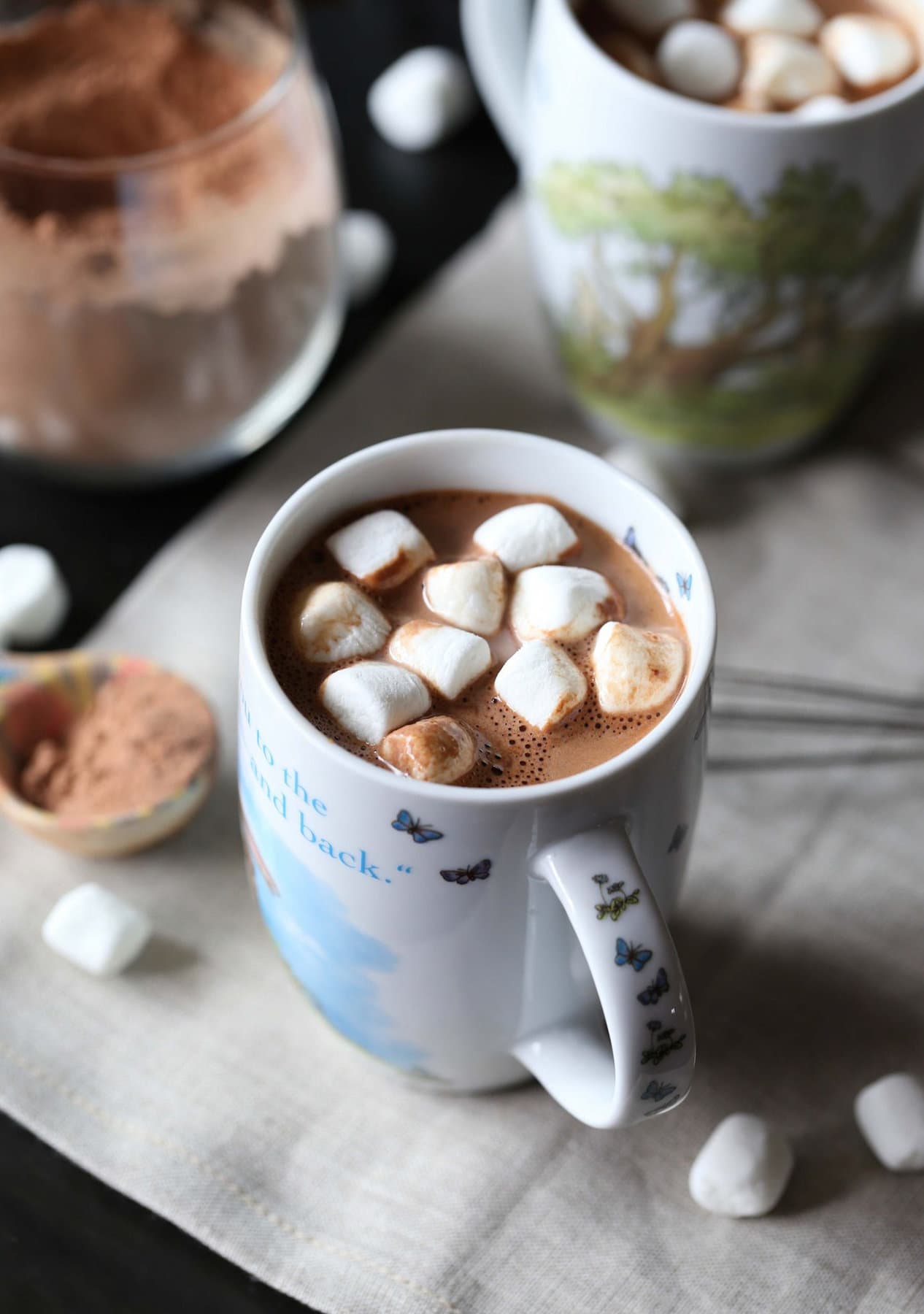 A mug of homemade hot cocoa topped with mini marshmallows, next to a jar of hot chocolate mix and a second mug of hot cocoa in the background.