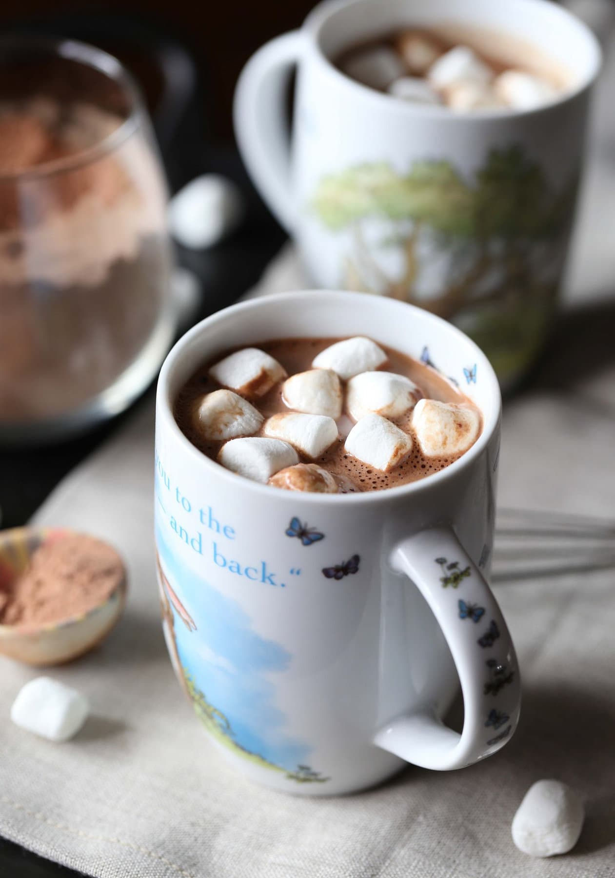A mug of homemade hot cocoa topped with mini marshmallows, next to a jar of hot chocolate mix and a second mug of hot cocoa in the background.