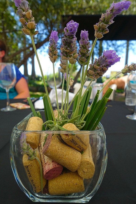 A glass vase on a table containing wine corks and purple flowers