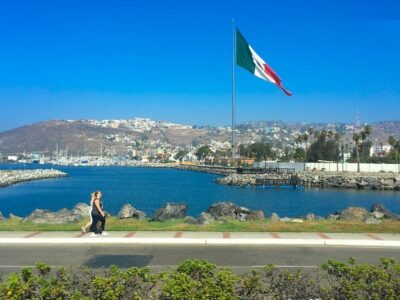 A lake in Ensenada with a large Mexican flag waving behind it and two people walking in front of it