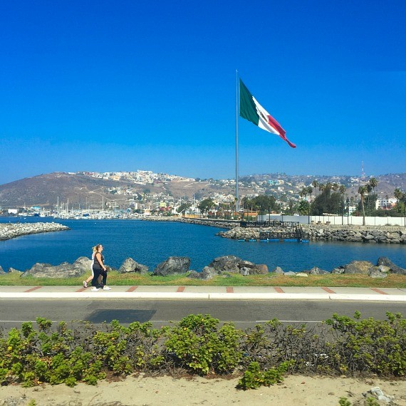 A lake in Ensenada with a large Mexican flag waving behind it and two people walking in front of it