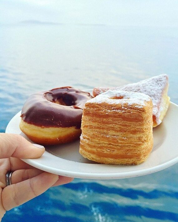 A chocolate frosted doughnut and two other pastries on a plate with ocean water in the background