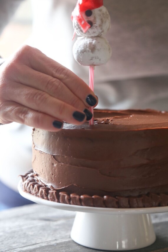 A hand gently pressing the bottom of a cake pop stick topped with a snowman into the top of a frosted chocolate cake.