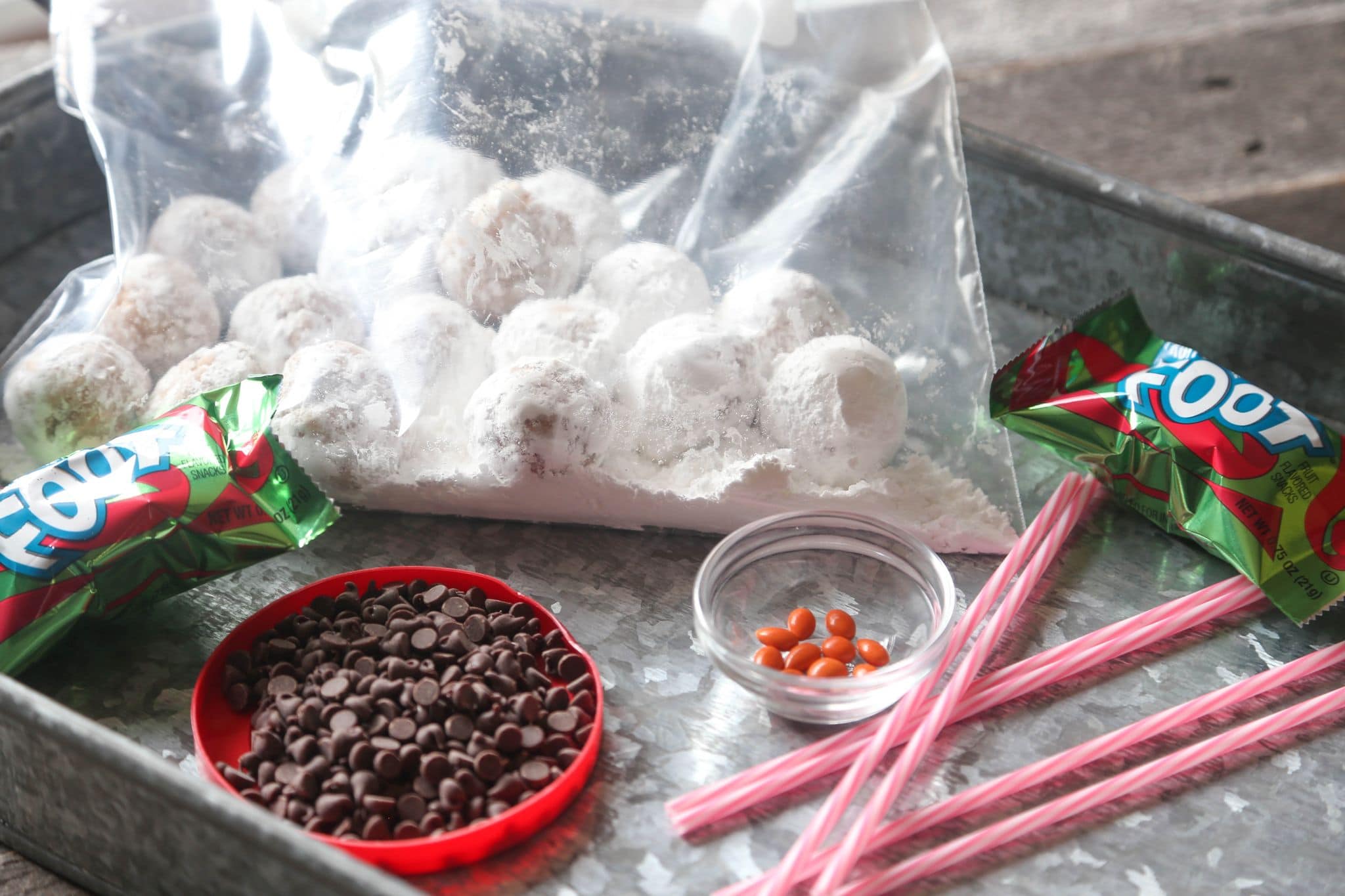 Supplies needed to decorate a snowman cake, including a bag of powdered donut holes, chocolate chips, and cake pop sticks.