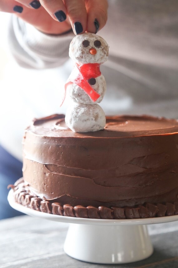 A snowman cake pop being pressed into the top of a frosted chocolate cake.