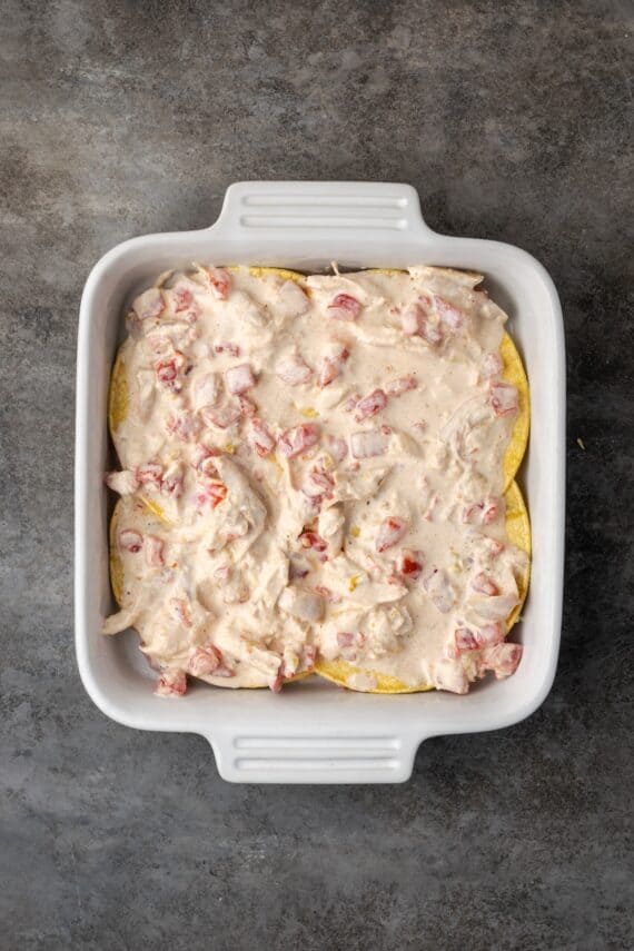 Salsa verde chicken layered over corn tortillas in a square ceramic baking dish.