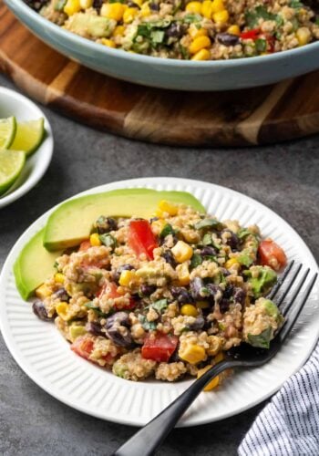 A serving of Southwestern quinoa salad next to avocado slices on a white plate with a fork.