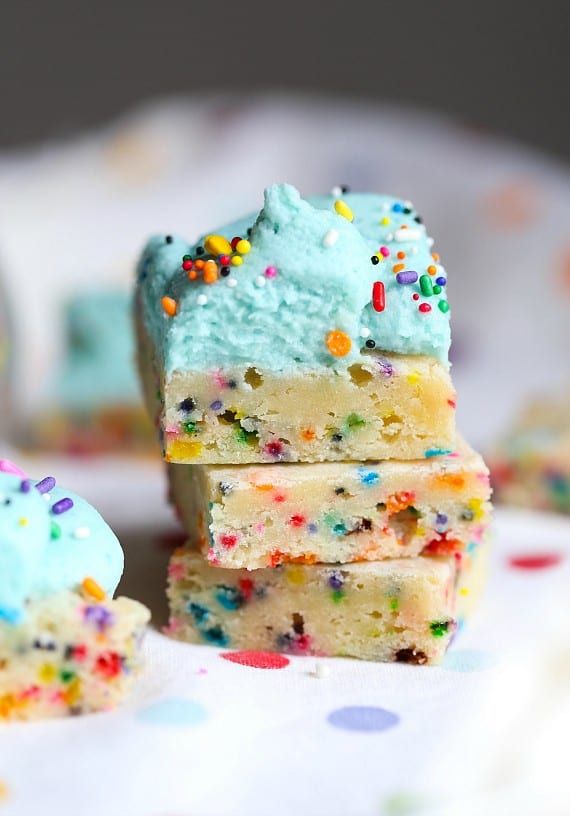 Unicorn bars are a fun, easy dessert! Buttery, soft-baked funfetti cookie bars loaded with rainbow sprinkles and frosted with homemade icing. Three unicorn bars stacked with the top bar decorated with fluffy blue frosting and sprinkles.
