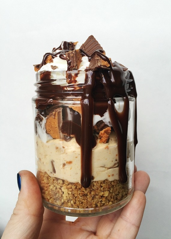 A hand holding up a no bake Butterfinger cheesecake cup loaded with Butterfinger candies and drizzled with chocolate sauce.