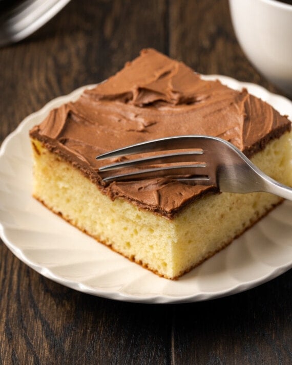 A fork cuts into the corner of a slice of frosted sour cream coffee cake on a white plate.