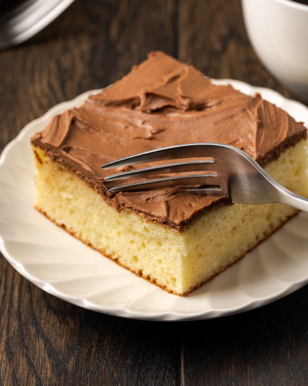 A fork cuts into the corner of a slice of frosted sour cream cake on a white plate.
