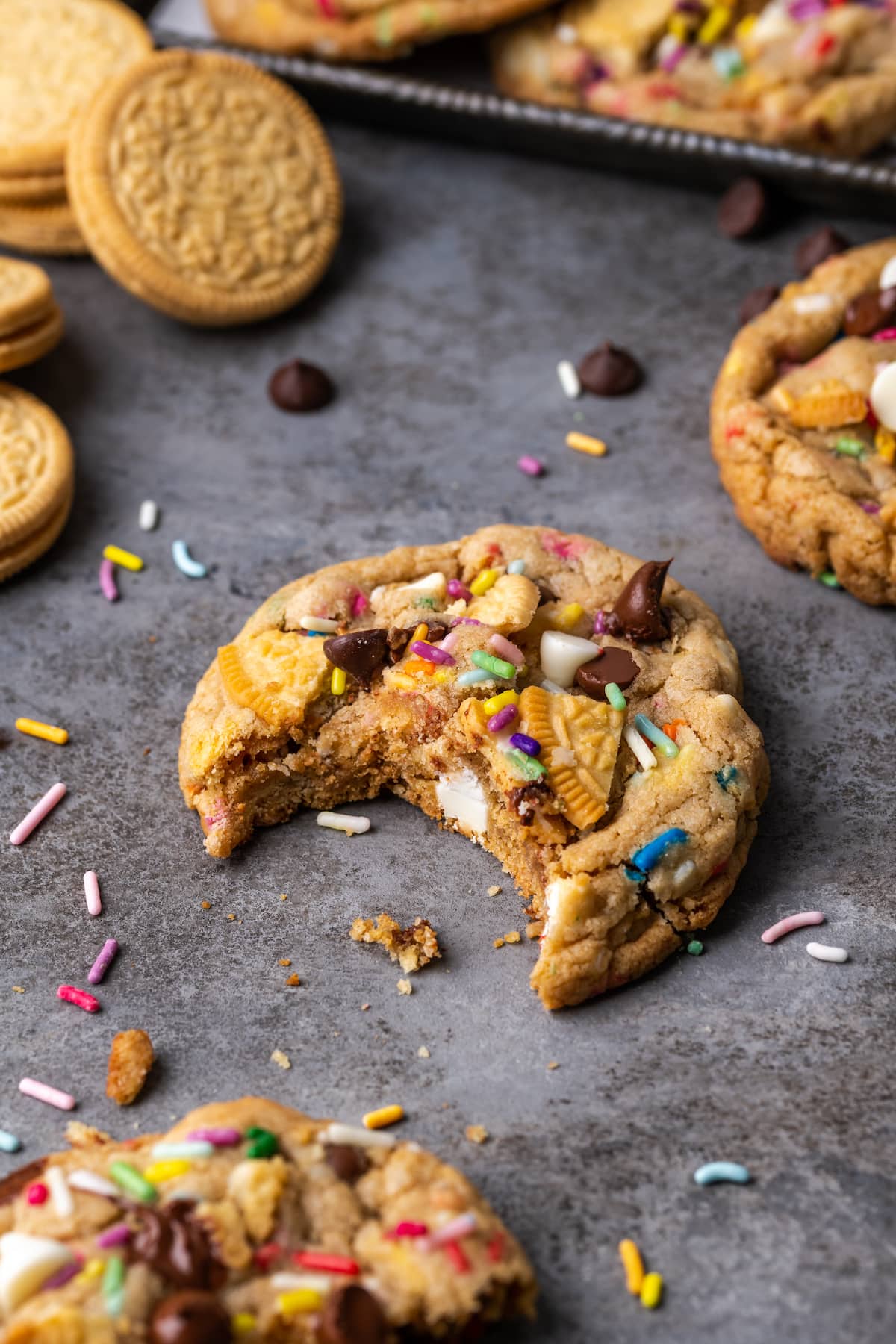 A birthday cake chocolate chip cookie with a bite missing on a countertop, surrounded by more cookies.