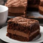 A slice of frosted fudge cake on a white plate with the rest of the cake in the background.