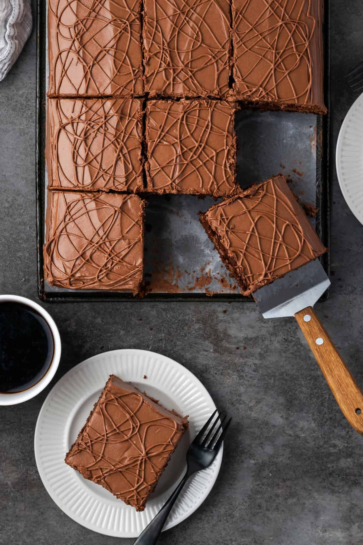 Overhead view of a frosted fudge cake cut into slices, with one slice being served from the cake pan, next to another slice served on a white plate with a fork.