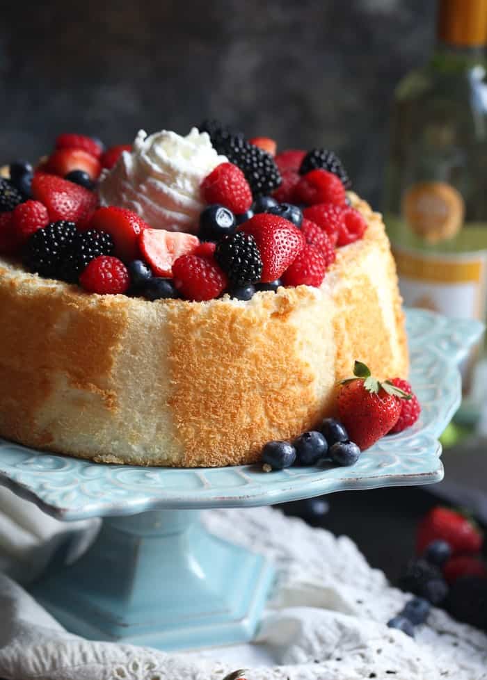 This light, airy angel food cake recipe is the perfect resting place for a juicy wine-soaked berry topping. An easy, elegant summer dessert! Angel food cake topped with macerated berries and whipped cream on a cake stand.