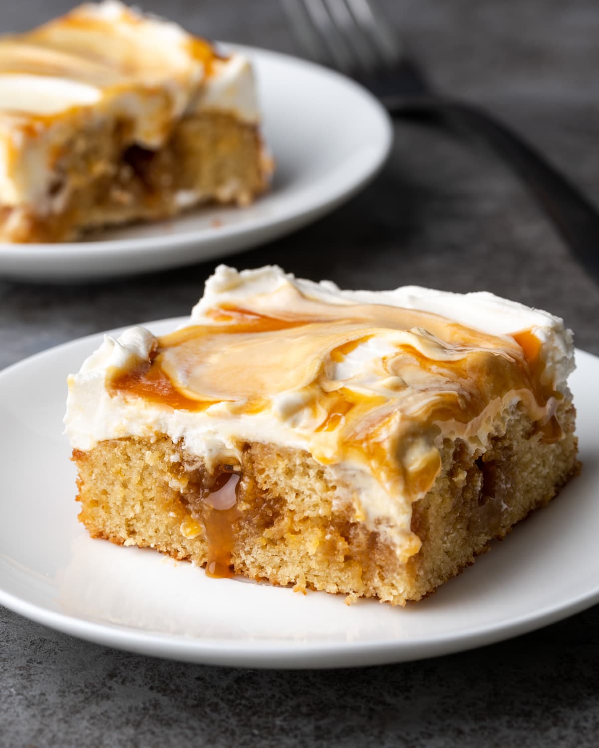 A slice of frosted caramel cake on a white plate, with a second serving in the background.