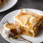 A slice of frosted caramel cake on a white plate with a forkful missing.