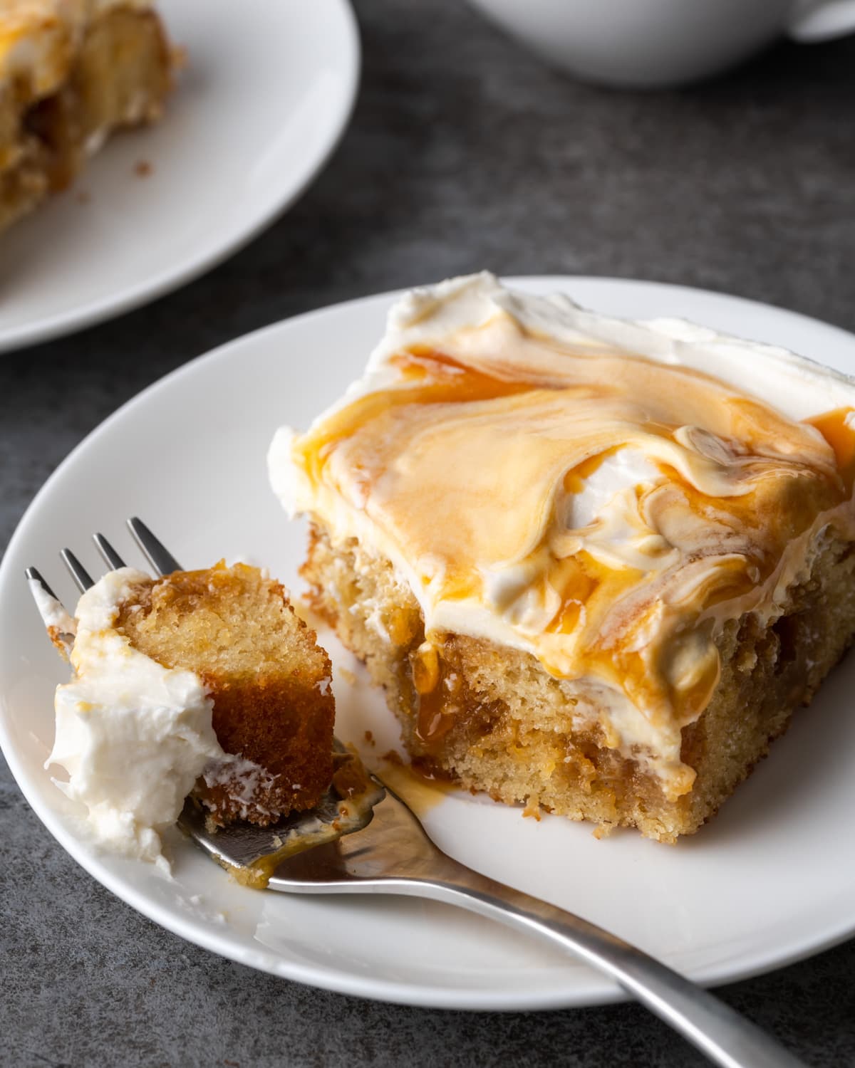 A slice of frosted caramel cake on a white plate with a forkful missing.