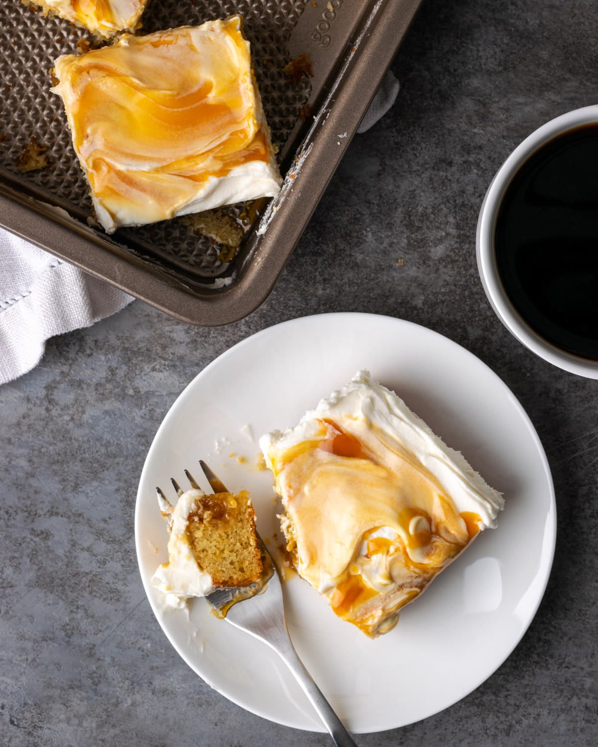 Top view of a slice of caramel cake on a plate next to a forkful, next to more slices in a pan and a cup of coffee.