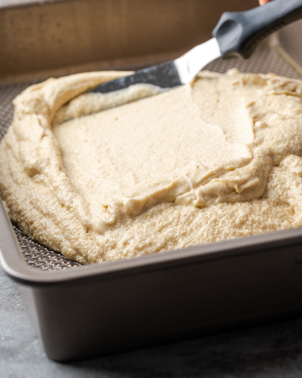 Vanilla cake batter is spread into a baking pan with an offset spatula.