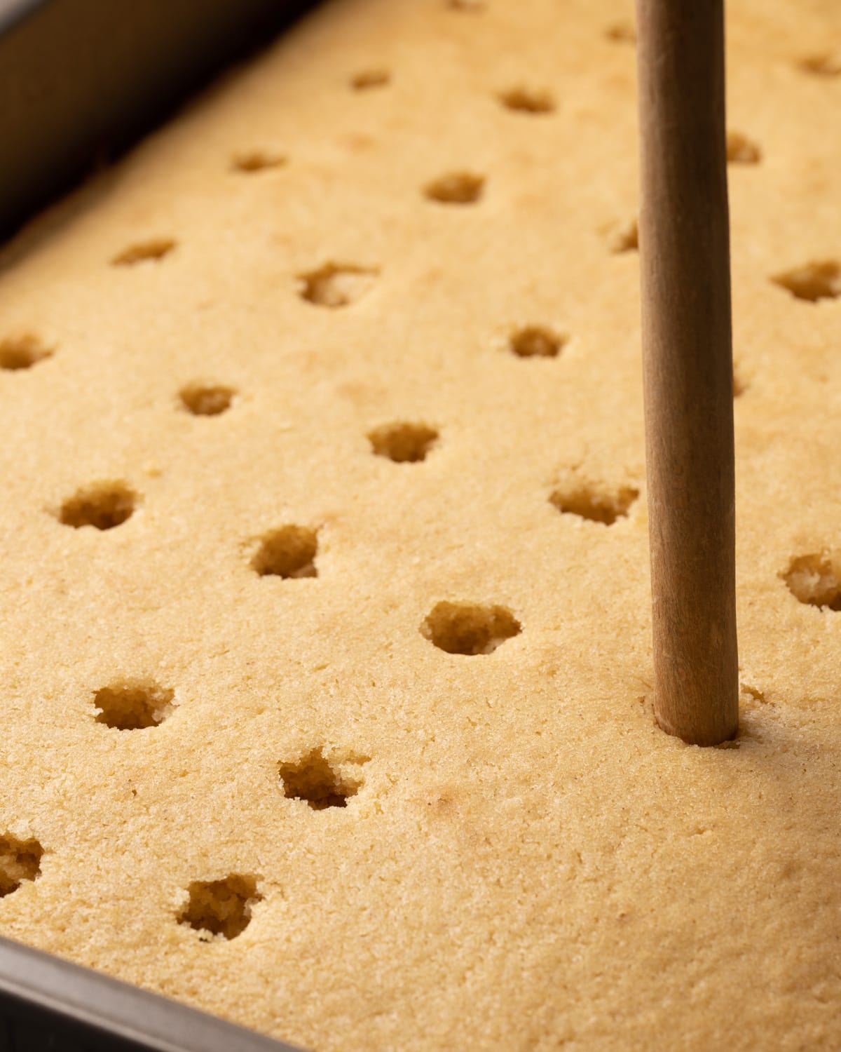 The back of a wooden spoon is used to poke holes in a baked vanilla cake while it's still warm in the pan.