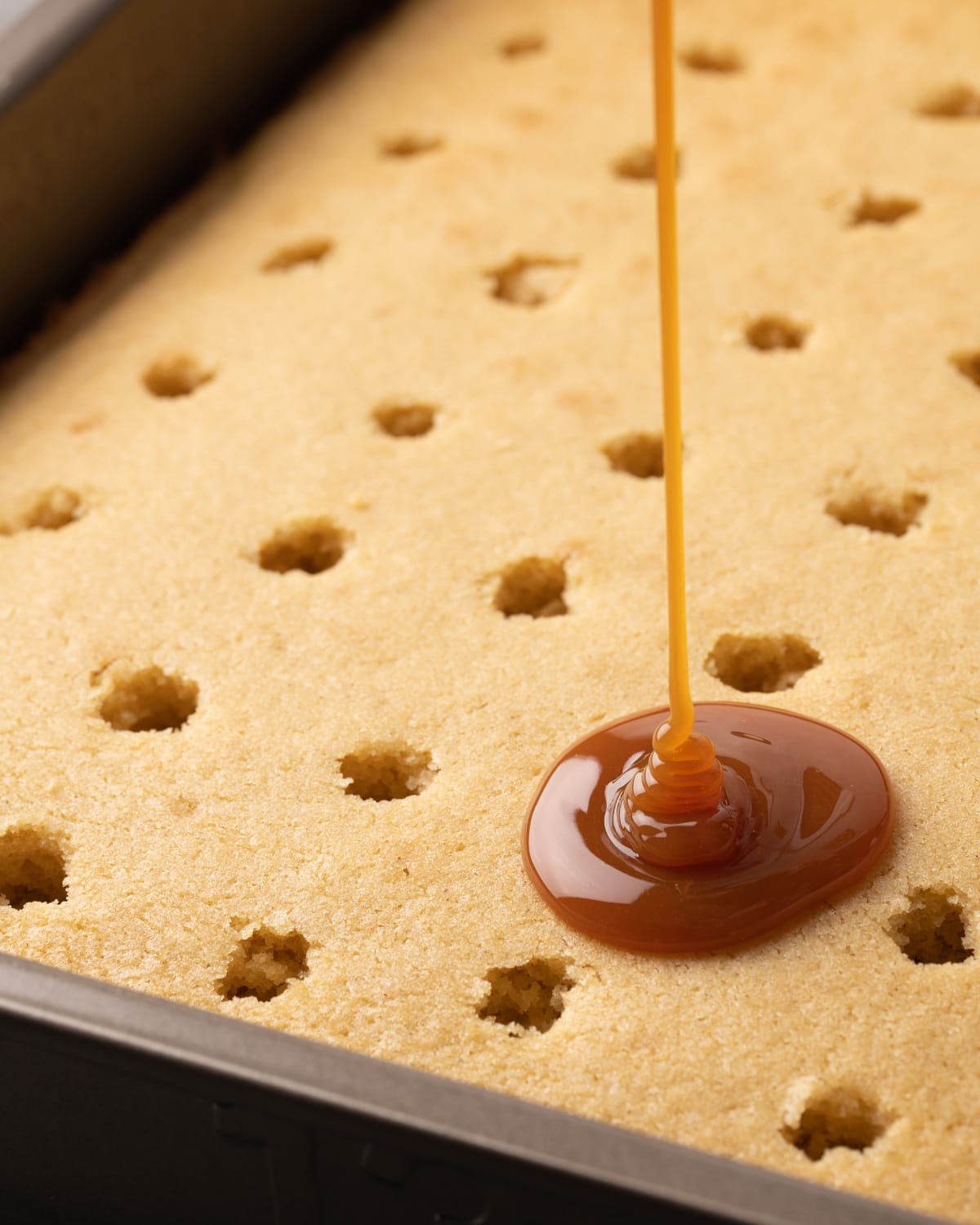 A stream of caramel is drizzled onto a vanilla cake filled with holes.