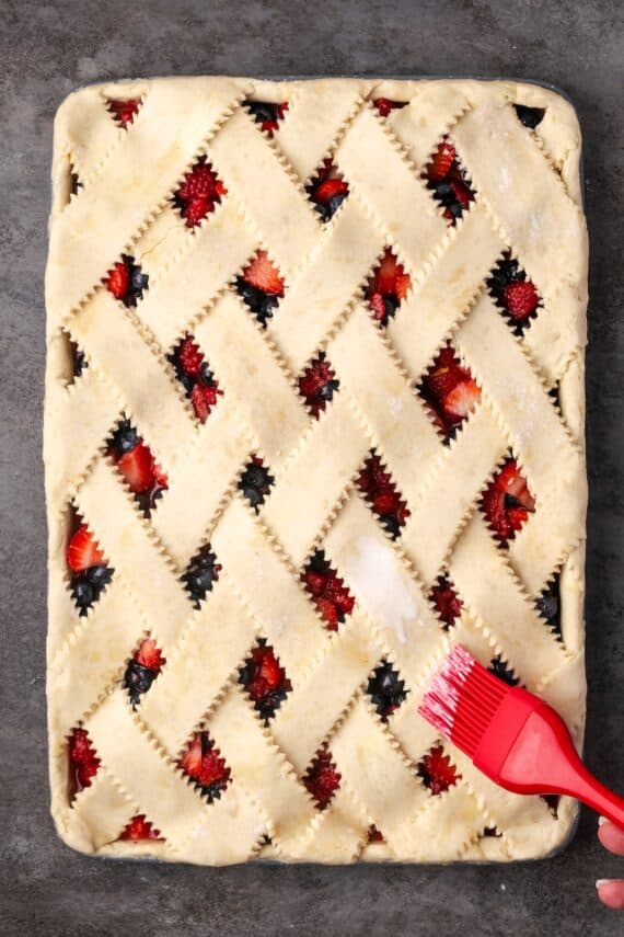 A rubber basting brush coating the lattice crust on top of a berry slab pie with an egg wash.