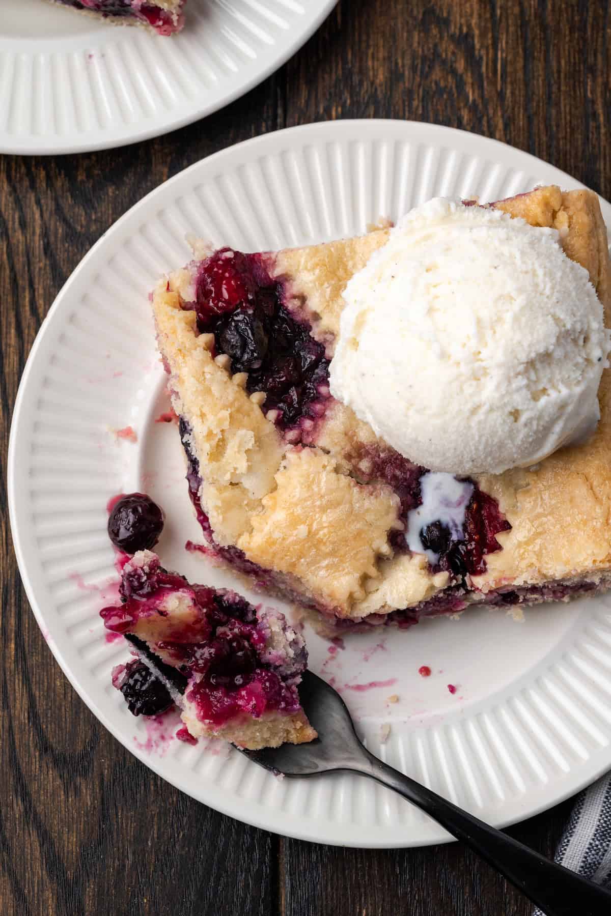 Overhead view of a slice of berry slab pie topped with a scoop of vanilla ice cream, next to a forkful of pie on a plate.
