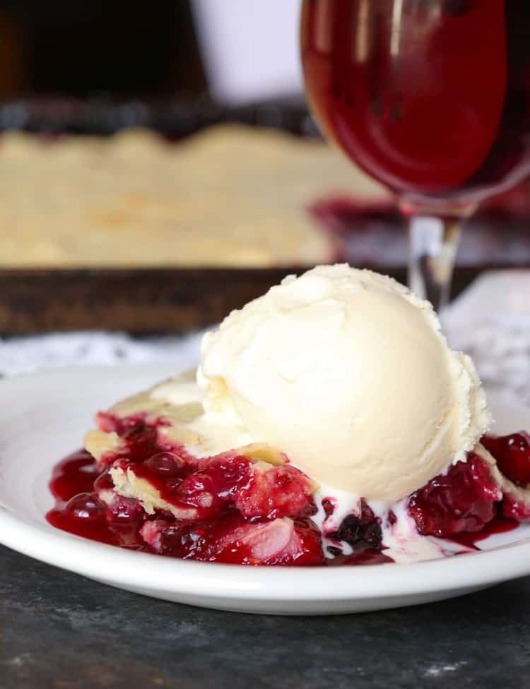A slice of Berry Slab Pie on a white plate topped with a scoop of vanilla ice cream.