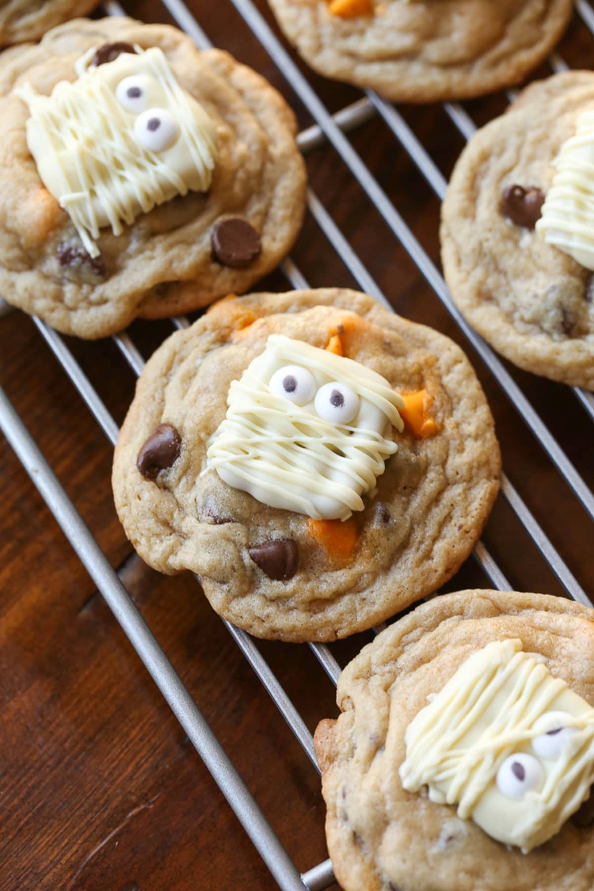 Chocolate chip cookies on a wire cooling rack with orange chips topped with a white chocolate pretzel decorated like a mummy.