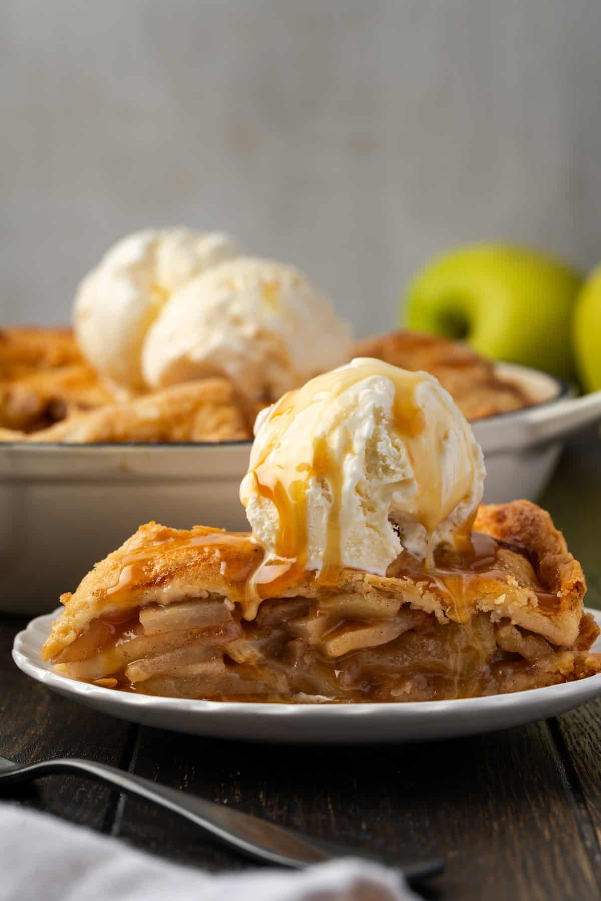 A slice of skillet apple pie topped with a scoop of vanilla ice cream on a white plate, with the rest of the pie in a skillet in the background.