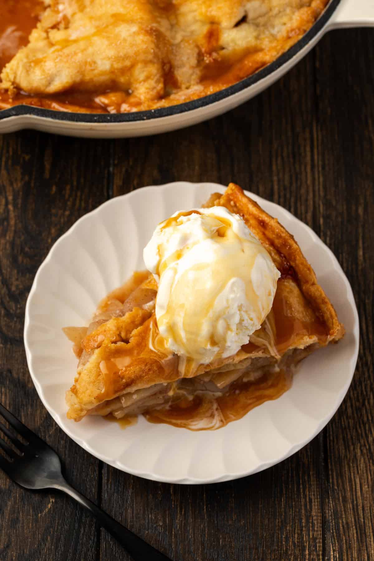 Overhead view of a slice of skillet apple pie topped with a scoop of vanilla ice cream on a white plate, next to the rest of the pie in a skillet.