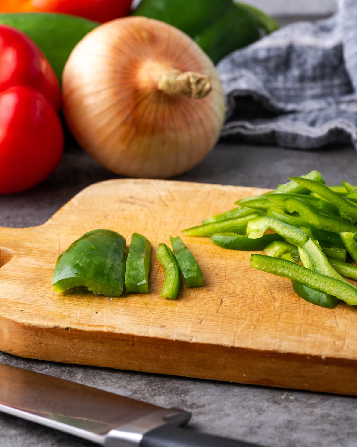 Chopped green bell peppers on a wooden cutting board, with an onion and tomatoes in the background.
