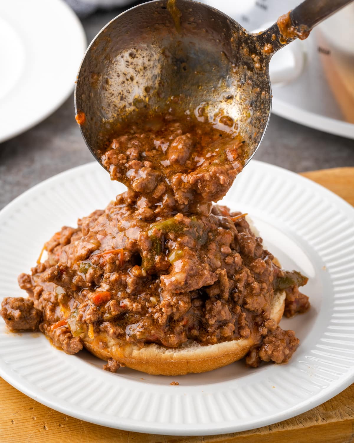 A ladle scoops Philly Cheesesteak Sloppy Joes over one half of a hamburger bun on a plate.