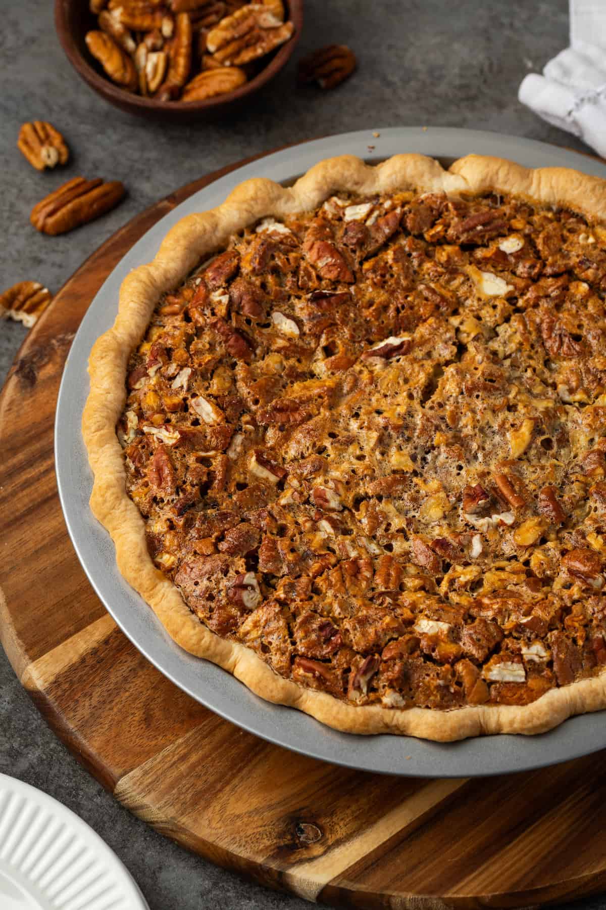 A whole cheesecake pecan pie in a pie plate on a round wooden platter, with a bowl of pecans in the background.