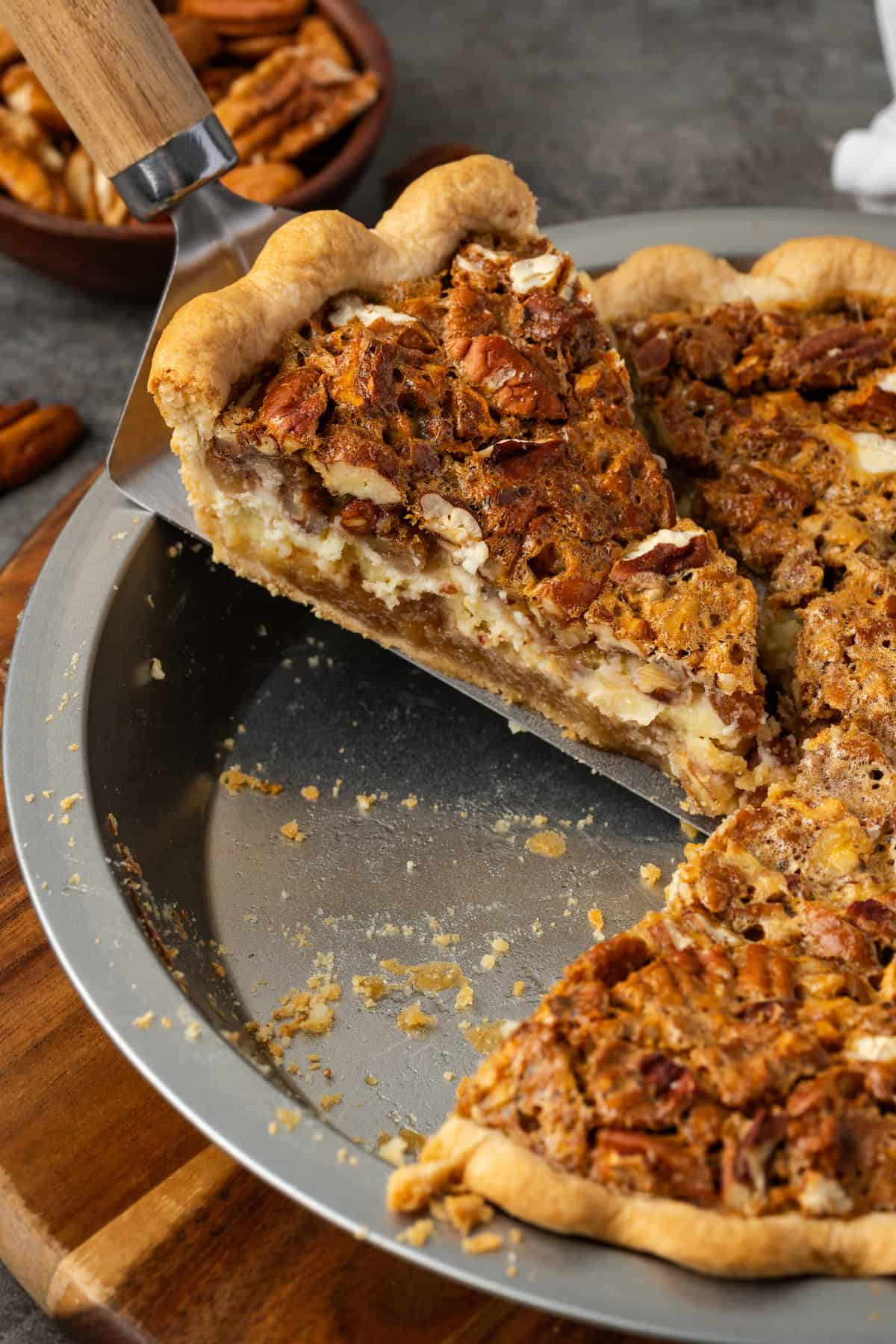 Close up of a slice of cheesecake pecan pie being lifted from the rest of the pie in a pie plate.