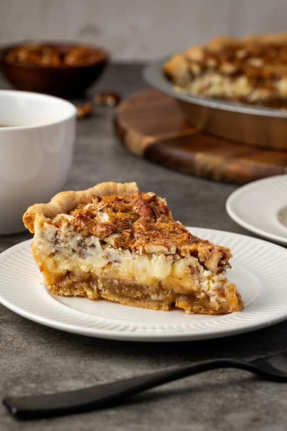 A slice of cheesecake pecan pie on a white plate next to a cup of coffee, with the rest of the pie in the background.