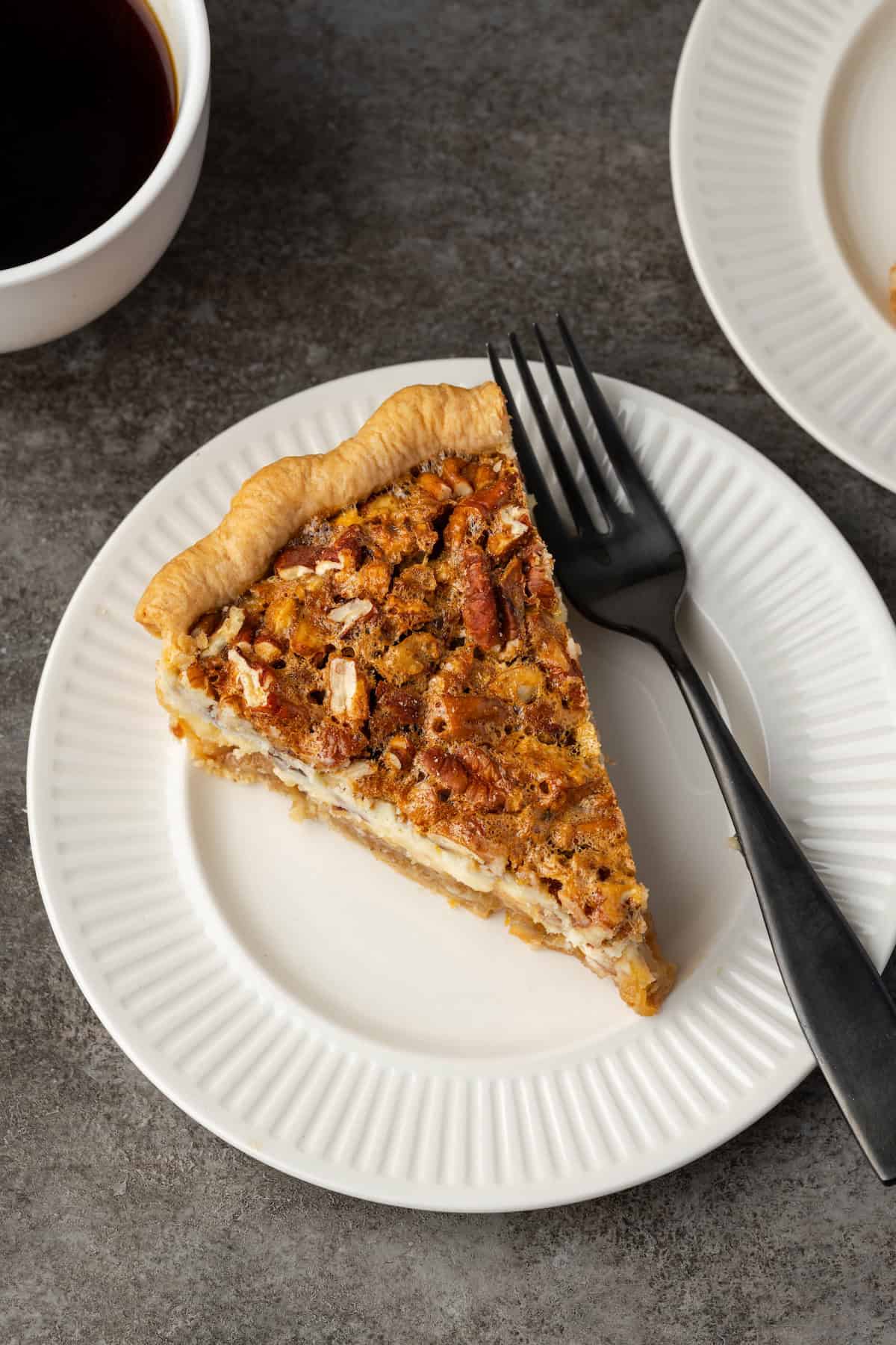 Overhead view of a slice of cheesecake pecan pie on a white plate next to a fork.