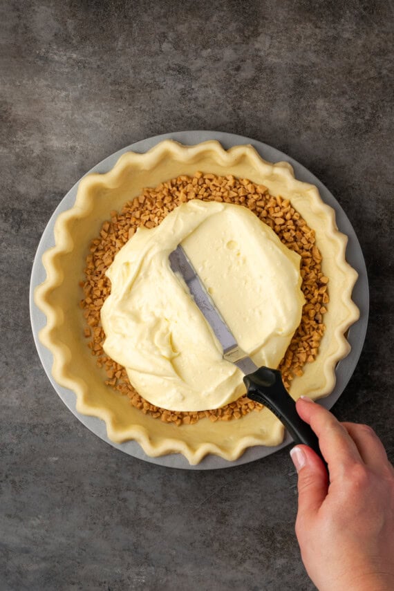 A hand using an offset spatula to spread cheesecake batter over toffee bits in a pie crust.