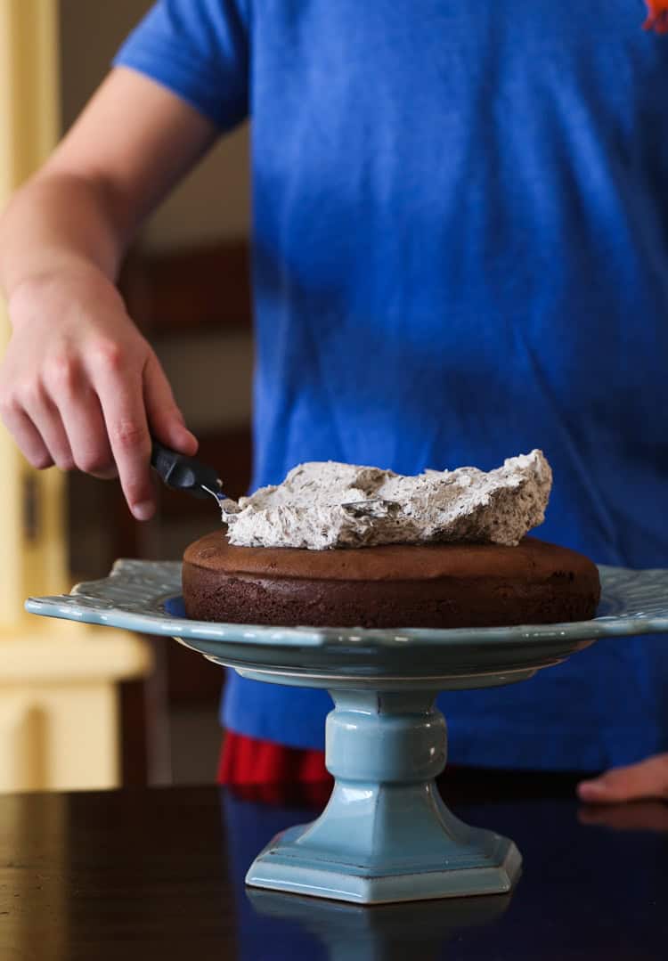 A Boy Spreading Cookies and Cream Buttercream Over a Chocolate Cake on a Light Blue Stand