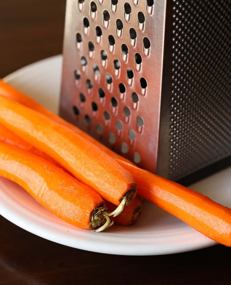 The best carrot cake recipe ever! Layers of moist cake filled with carrots, pineapple, and coconut, covered with rich cream cheese frosting. Peeled carrots next to a box grater.