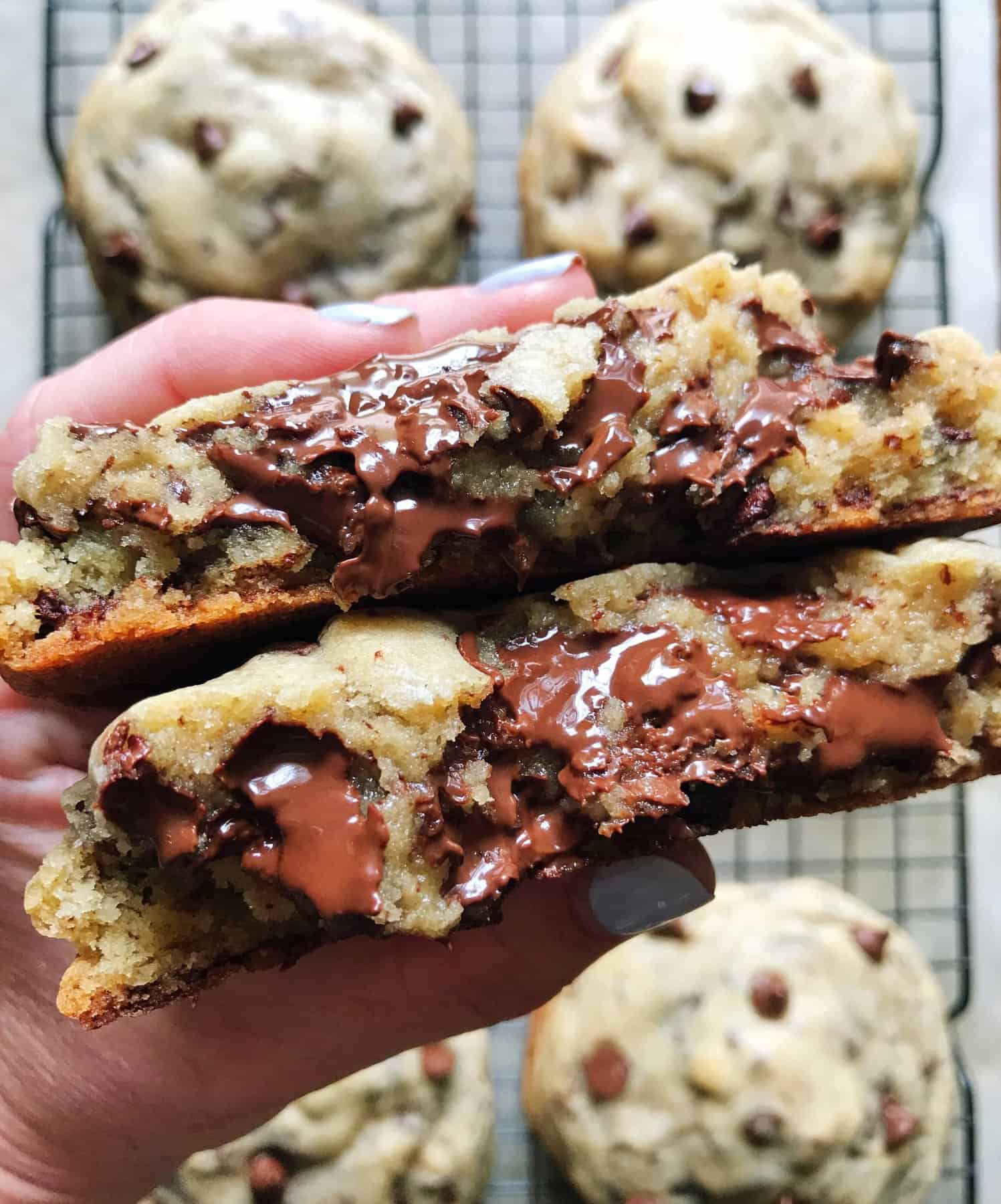 Two Gooey Chocolate Chip Cookies Being Held Above More Cookies on a Cooling Rack