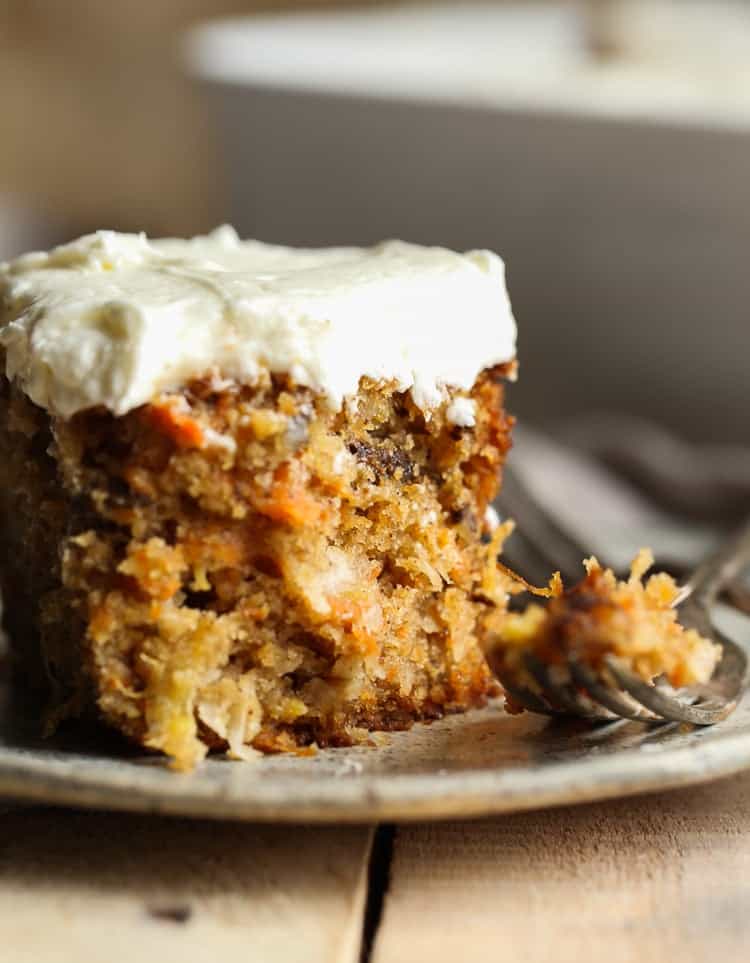A moist and fluffy slice of frosted carrot cake on a plate next to a fork with a fresh bite of cake