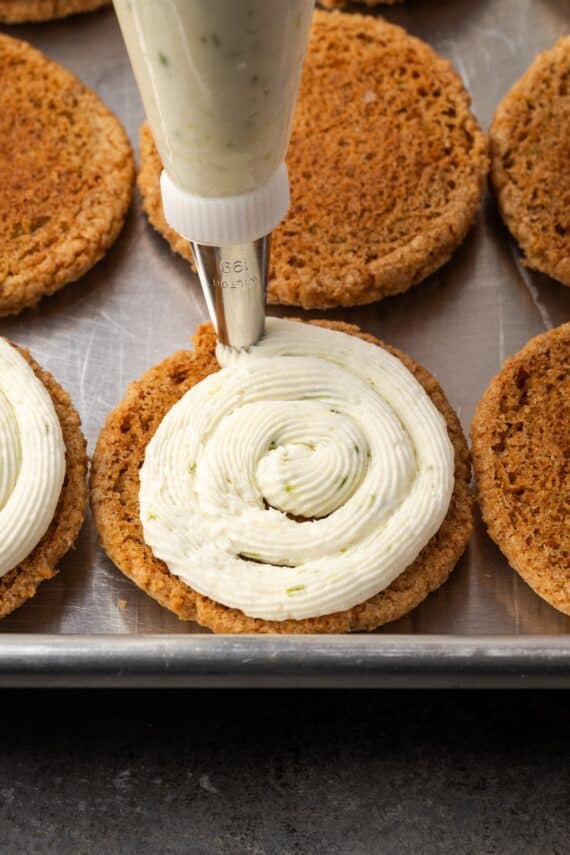 A piping tip pipes key lime buttercream onto a graham cookie on a baking sheet, surrounded by more unfrosted cookies in the background.