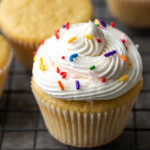 A vanilla cupcake topped with a swirl of buttercream frosting and rainbow sprinkles, next to more cupcakes on a wire rack.