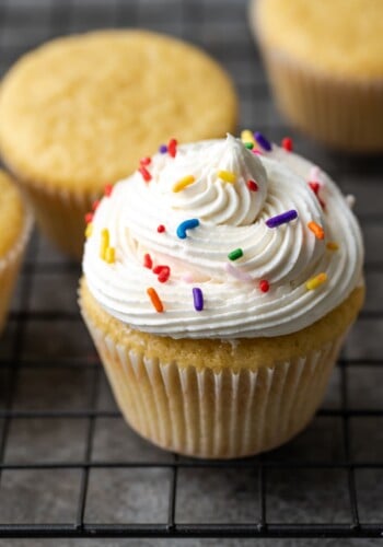 A vanilla cupcake topped with a swirl of buttercream frosting and rainbow sprinkles, next to more cupcakes on a wire rack.